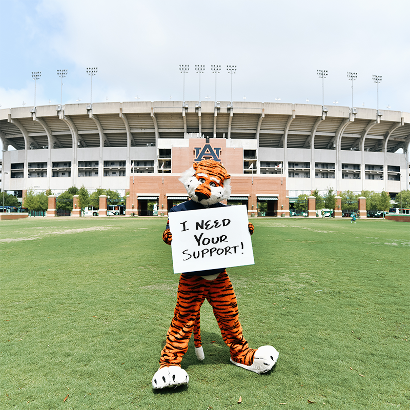 Aubie standing in front of Jordan-Hare stadium holding a sign that says I need your support