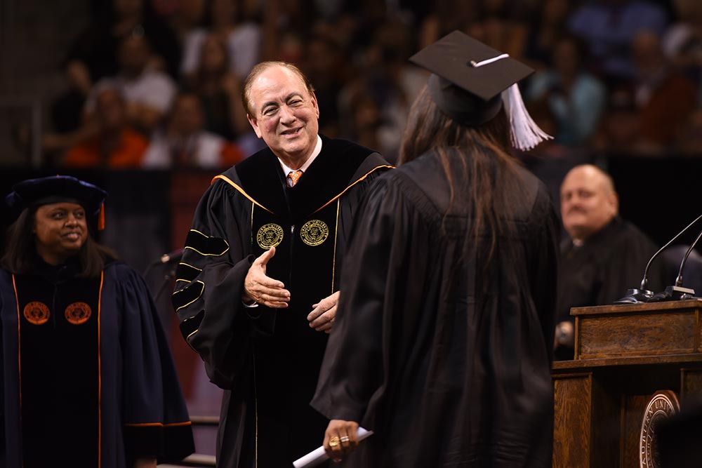 President Jay Gogue smiles on stage during graduation as he prepares to shake hands with a student.