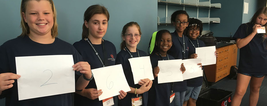 Six girl students holding up a piece of paper with a number on it.
