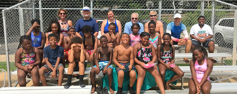 Older group of boys and girls in swim gear sit in bleachers along with swim instructors to pose for photo