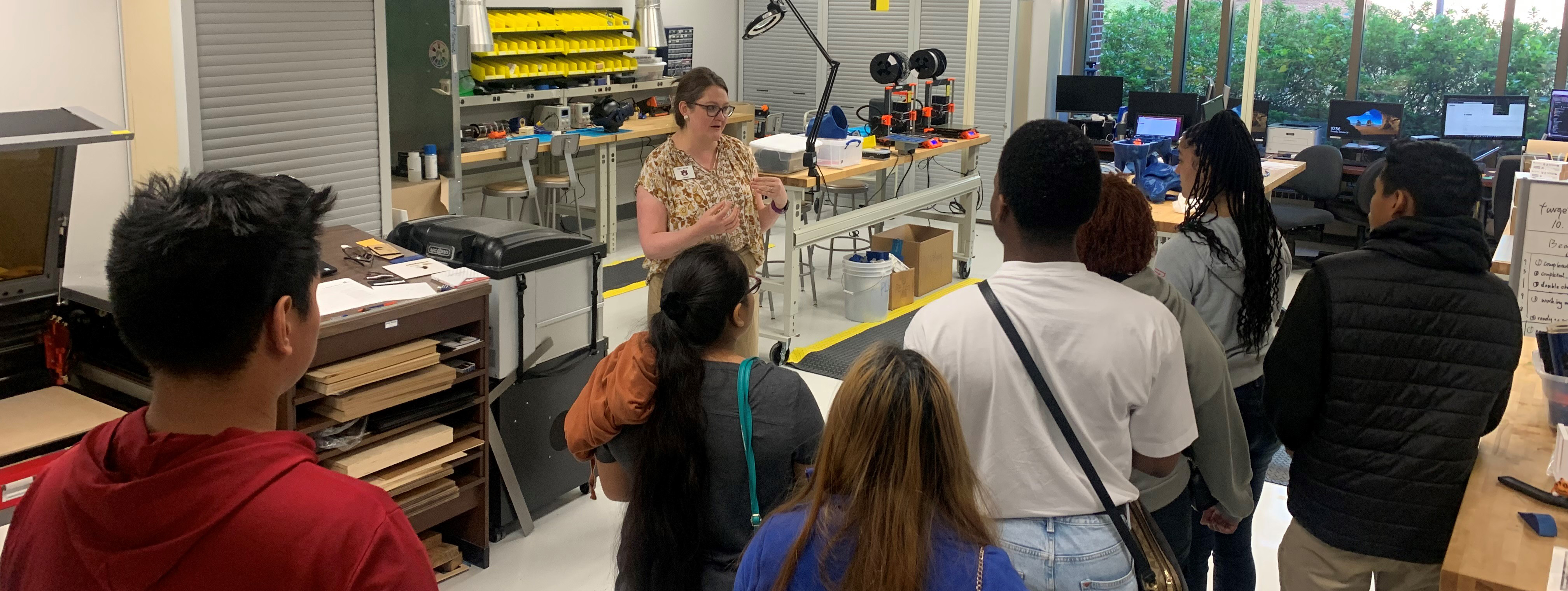 A female presenter speaks to a group of male and female students in a makers space lab