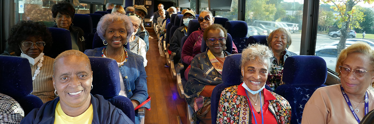 A group of women and men sit on a bus with blue seats smiling.