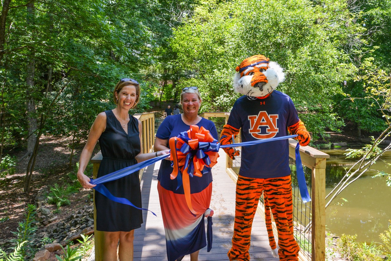 Ashley Underwood from COSAM Development, Arboretum Director Morgan Beadles, and of course Aubie stand in front of the new bridge to celebrate the ribbon cutting. 