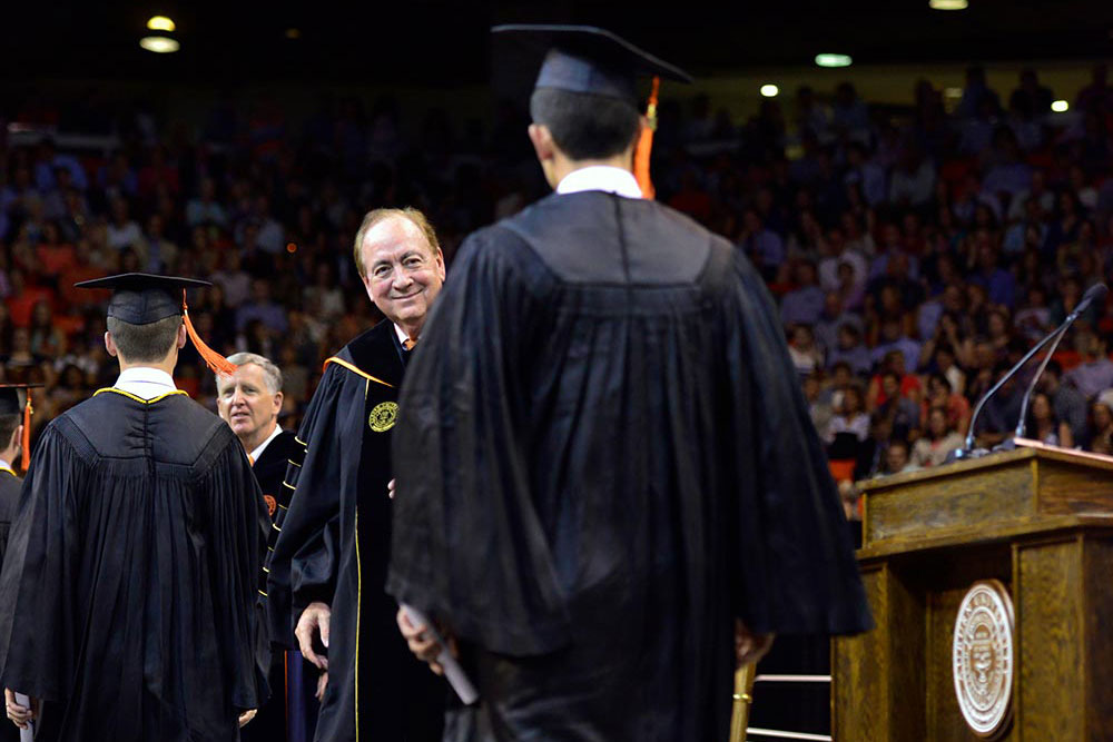 President Jay Gogue stands on stage during a commencement ceremony as he prepares to shake hands with a new graduate.