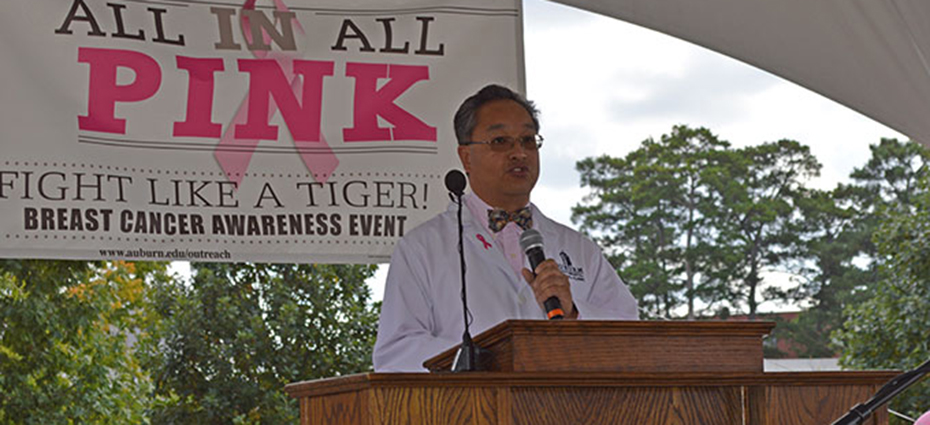 Dr. Fred Cam speaks at podium holding microphone with All in All pink banner behind him
