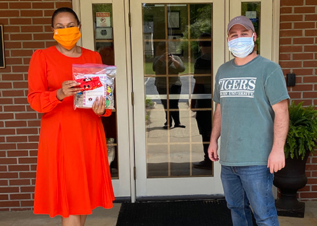 Woman in red dress holding bag of face masks and man in green Auburn shirt stand outside of brick building.