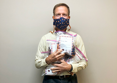 Man stands against wall wearing face mask and holding several ziploc bags of donated face masks.