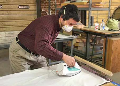 White man wearing a mask irons white fabric on a table