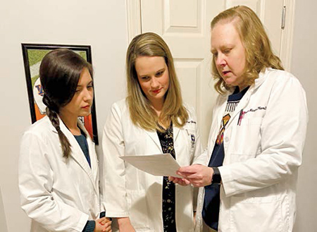 Three women wearing white coats stand together looking at papers