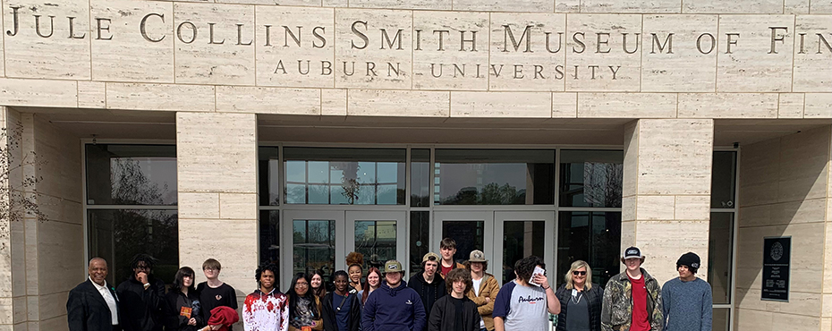 A large group of male and female students and teachers stand under a main entrance to a museum sign outside