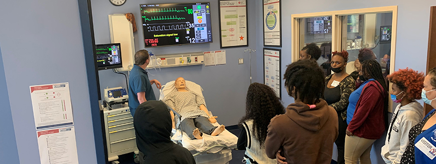 A small group of male and female students learn and observe medical assistance in a lab setting on a site tour and visit.