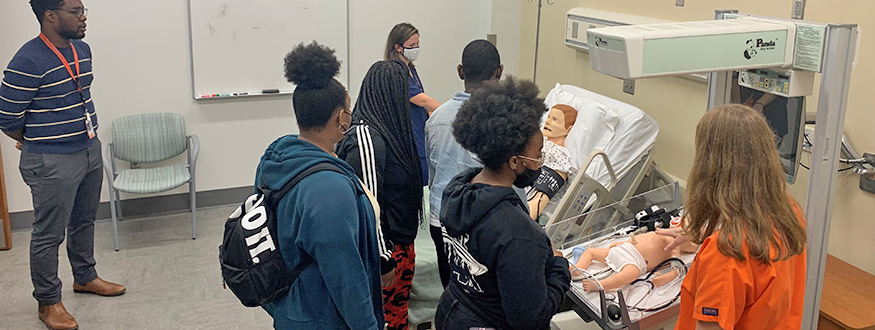 Students tour facility with a medical mannequin in a hospital beds and a mannequin baby in bassinet.