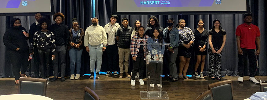 A group of male and female students stand in front of a podium and under a presentation screen