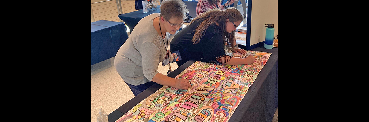 Two women use crayons to color in Outreach banner at New Faculty Orientation session.