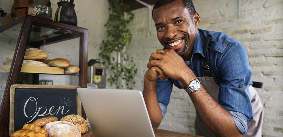 Man smiling and leaning on counter in bakery with laptop computer next to a sign that reads 'open'