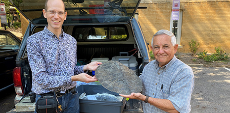 A picture containing 2 men standing outdoors in front of a blue truck holding a large fossilized turtle shell between them.