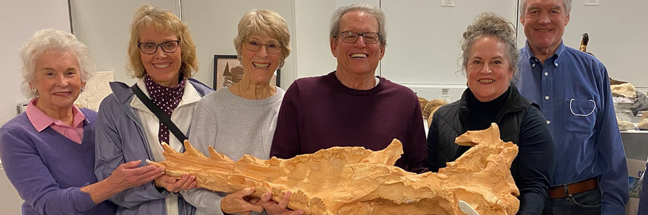 A group of older men and women stand side by side holding a long portion of a fossil.
