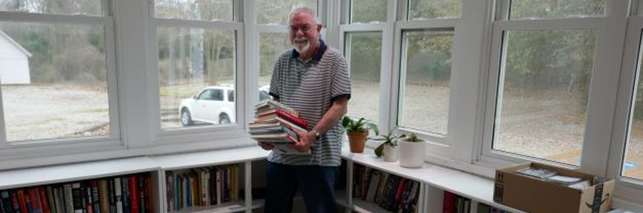 Man with white hair and beard holds a stack of books while standing in front of low bookcases under windows.