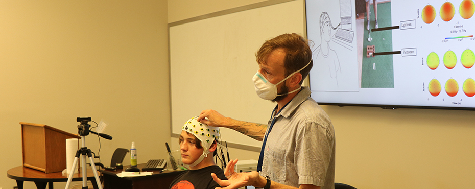 A male camper sits in a chair with an MRI cap on while an instructor speaks to the class.