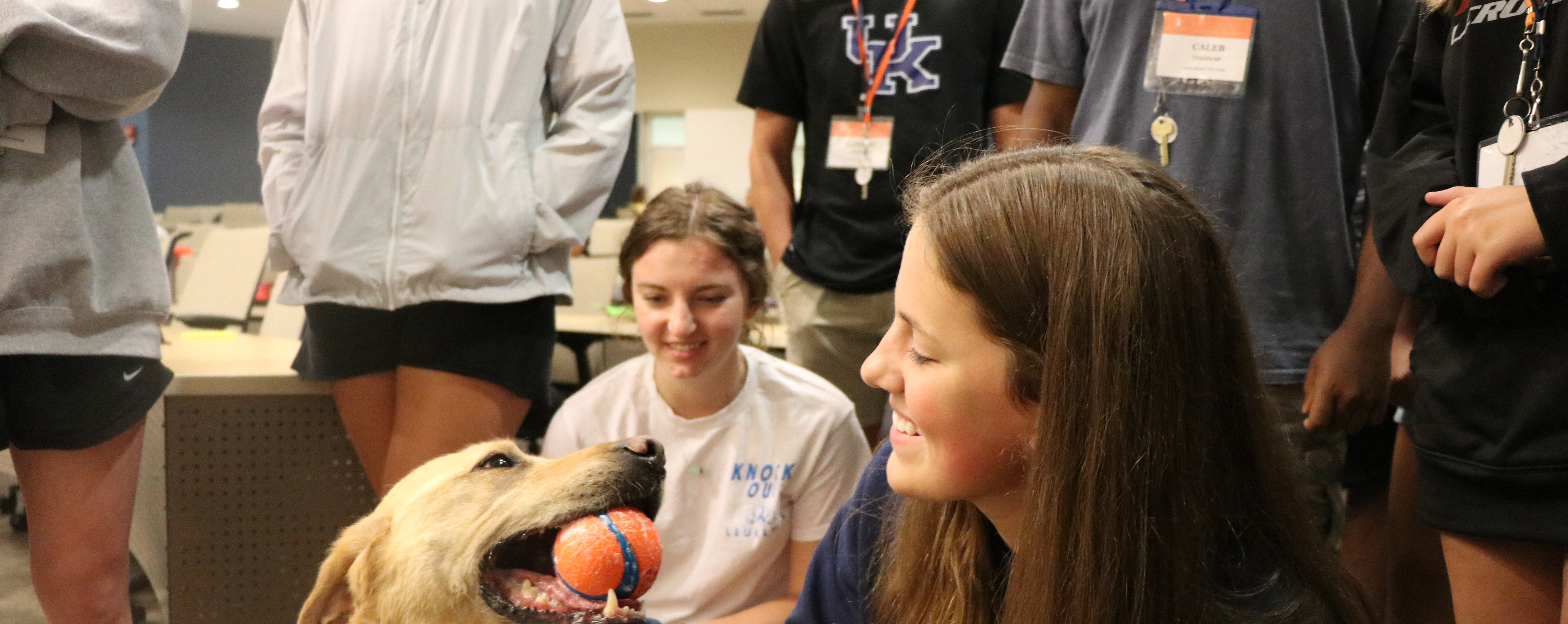 Campers play with a dog who is holding a ball in its mouth.