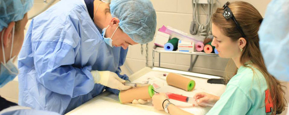 A student works with an instructor on learning how to draw blood using a pool noodle for practice.