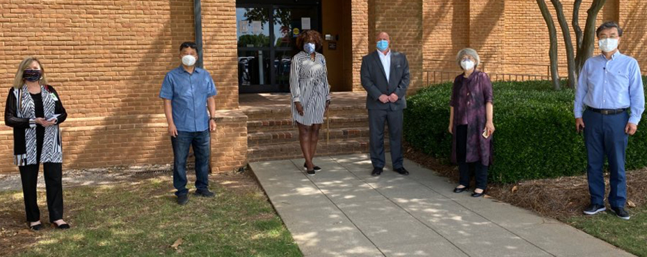 Six people stand outside of brick building wearing face masks.