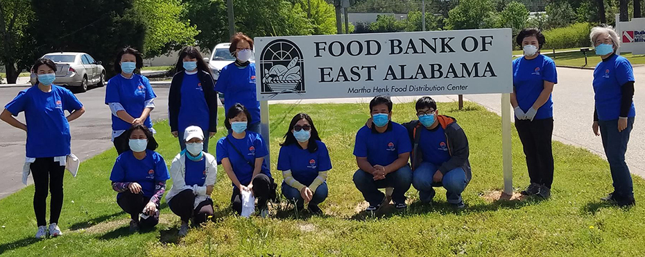 Thirteen Korea Corner volunteers wearing blue shirts pose for photo with the Food Bank of East Alabama sign.