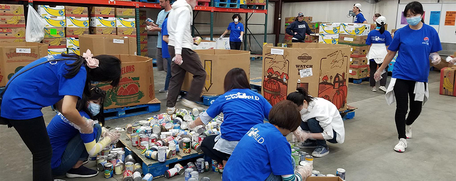 Volunteers in blue shirts and masks sort canned food into different boxes in large warehouse.